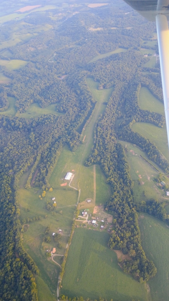 Aerial view of the Airborne Acres turf runway running through wooded hills, with the hangar and farm buildings in the foreground